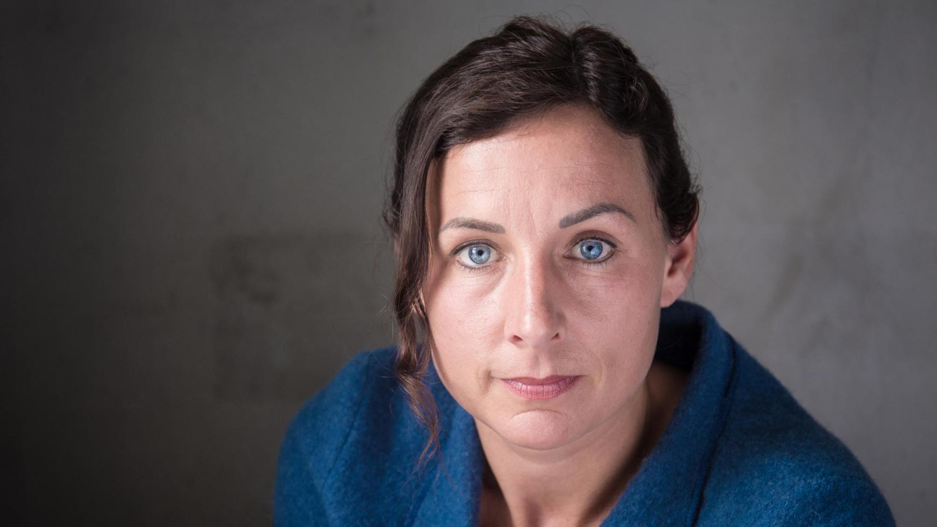 Juli Zeh sitzt in einer dunkelblauen Bluse vor einem dunklen Hintergrund und schaut in die Kamera. Juli Zeh sitzt in einer dunkelblauen Bluse vor einem dunklen Hintergrund und schaut in die Kamera.