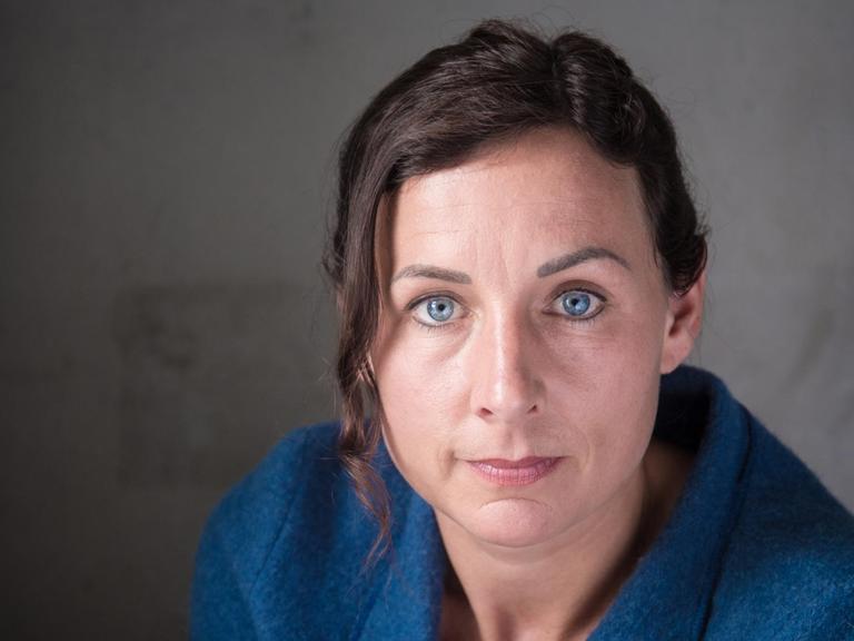 Juli Zeh sitzt in einer dunkelblauen Bluse vor einem dunklen Hintergrund und schaut in die Kamera.