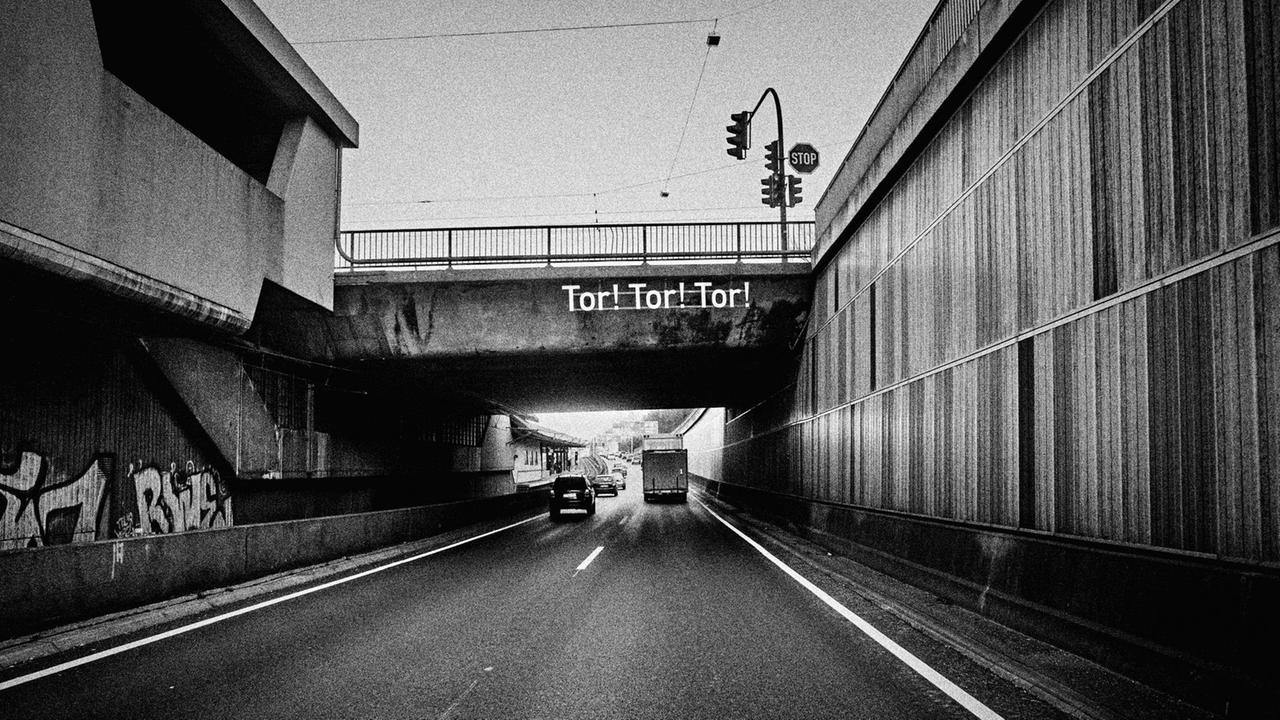 Ein Essener Stadtautobahn-Abschnitt in schwarzweiß fotografiert, umrahmt von grauen Wänden. Über ihn führt eine Brücke. An einer Stelle der Durchfahrt unter der Brücke ist der Slogan "Tor! Tor! Tor!" in großen Lettern zu lesen. Ein Essener Stadtautobahn-Abschnitt in schwarzweiß fotografiert, umrahmt von grauen Wänden. Über ihn führt eine Brücke. An einer Stelle der Durchfahrt unter der Brücke ist der Slogan "Tor! Tor! Tor!" in großen Lettern zu lesen.