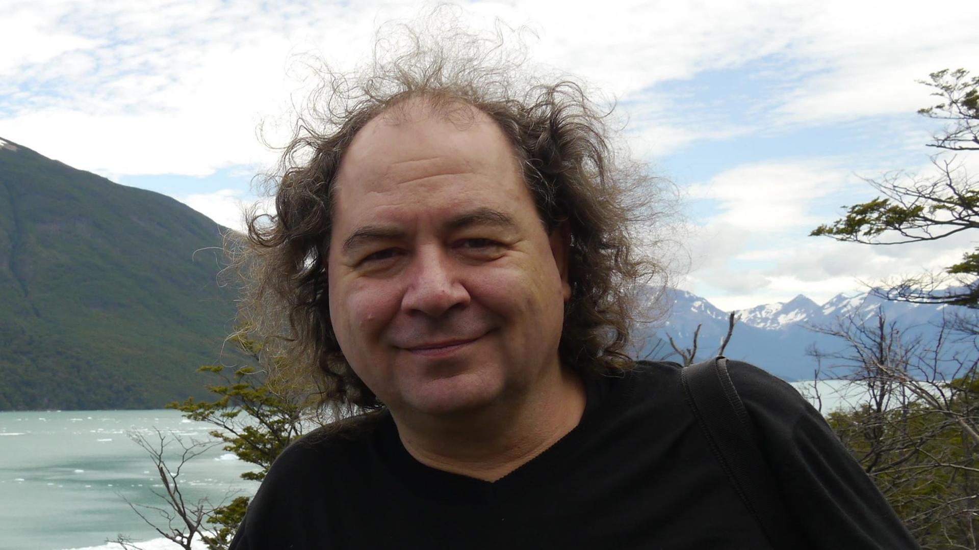 Ein lächelnder Mann mit lockigen Haaren steht vor einem gewaltigen Seen- und Bergpanorama. Ein lächelnder Mann mit lockigen Haaren steht vor einem gewaltigen Seen- und Bergpanorama.