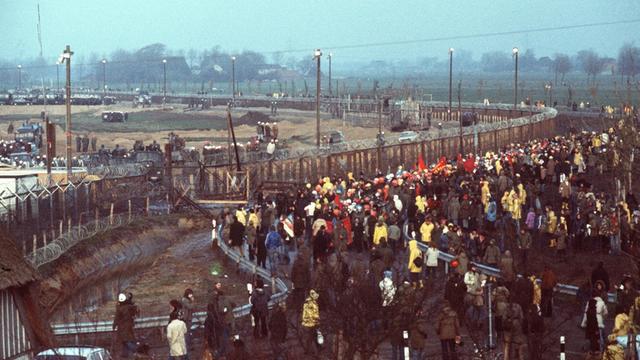 Demonstranten gegen den Kernkraftwerksbau in Brokdorf stehen mit Transparenten vor dem Baugelände, das mit Stacheldrahtzaun und Polizeiaufgebot abgesichert ist. Aufgenommen am 13. November 1976.