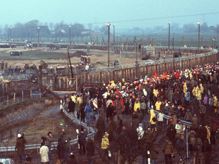 Demonstranten gegen den Kernkraftwerksbau in Brokdorf stehen mit Transparenten vor dem Baugelände, das mit Stacheldrahtzaun und Polizeiaufgebot abgesichert ist. Aufgenommen am 13. November 1976.
