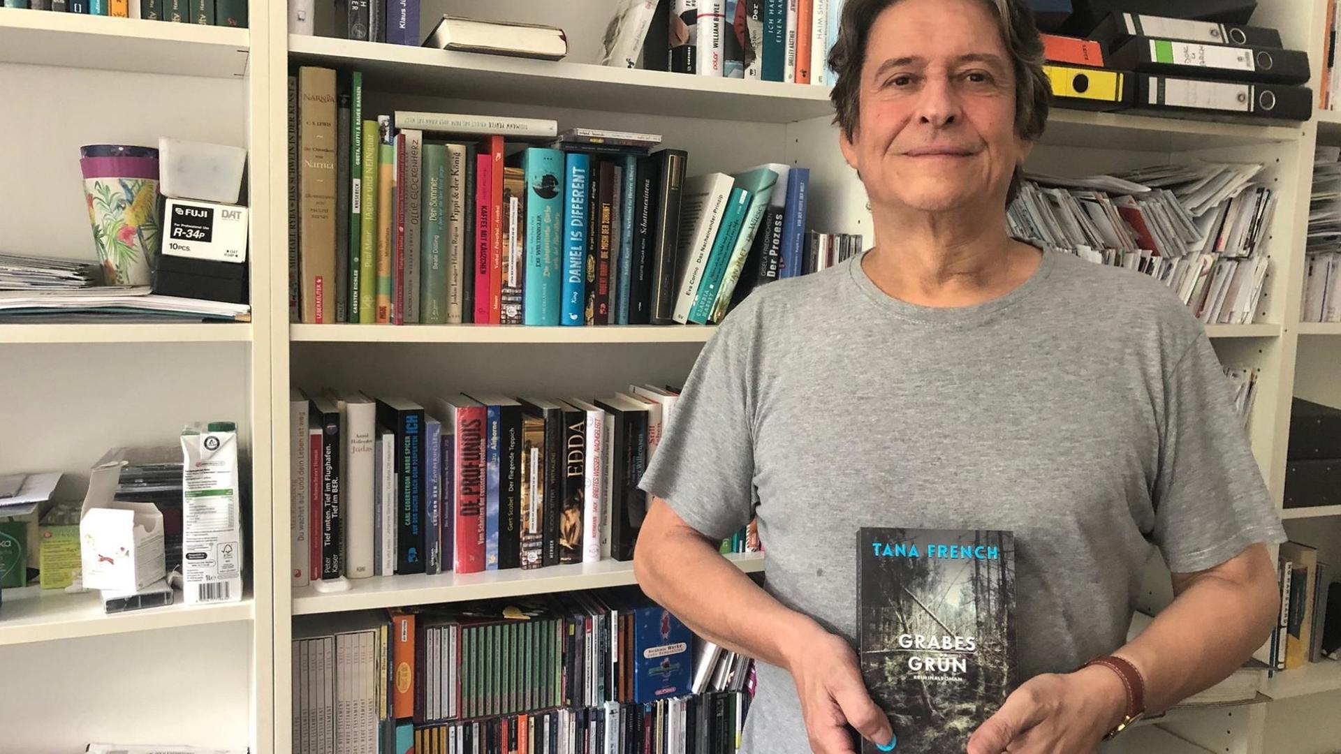 "Lesart"-Moderator Joachim Scholl steht vor einem Bücherregal und hält das Buch "Grabesgrün" von Tana French in der Hand.