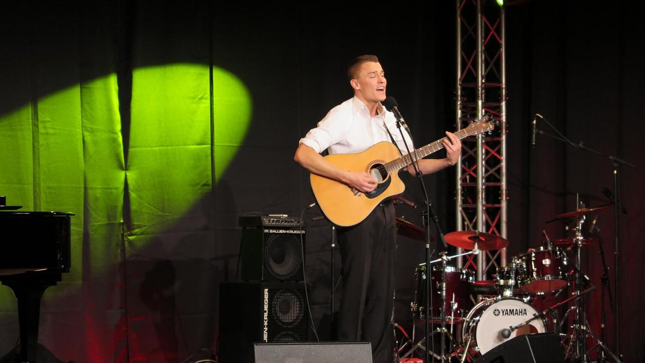 Alex Döring steht mit der Gitarre auf einer dunklen Bühne und begleitet seinen Gesang Alex Döring steht mit der Gitarre auf einer dunklen Bühne und begleitet seinen Gesang