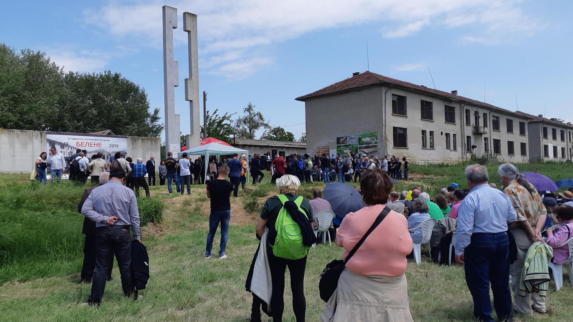 Etwa 100 Menschen versammeln sich auf einer Wiese, dahinter die Ruinen der Gefängnisbaracken Etwa 100 Menschen versammeln sich auf einer Wiese, dahinter die Ruinen der Gefängnisbaracken