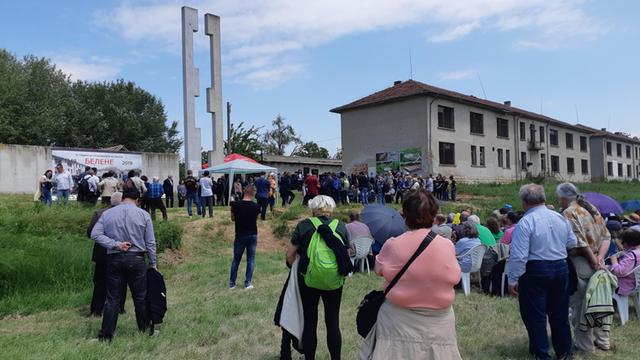Etwa 100 Menschen versammeln sich auf einer Wiese, dahinter die Ruinen der Gefängnisbaracken