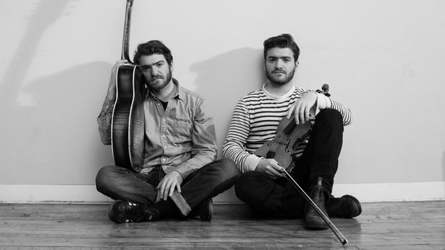 Links sitzt David Moss, mit einer Gitarre in der Hand, rechts sitzt Adam mit seiner Geige im Arm.