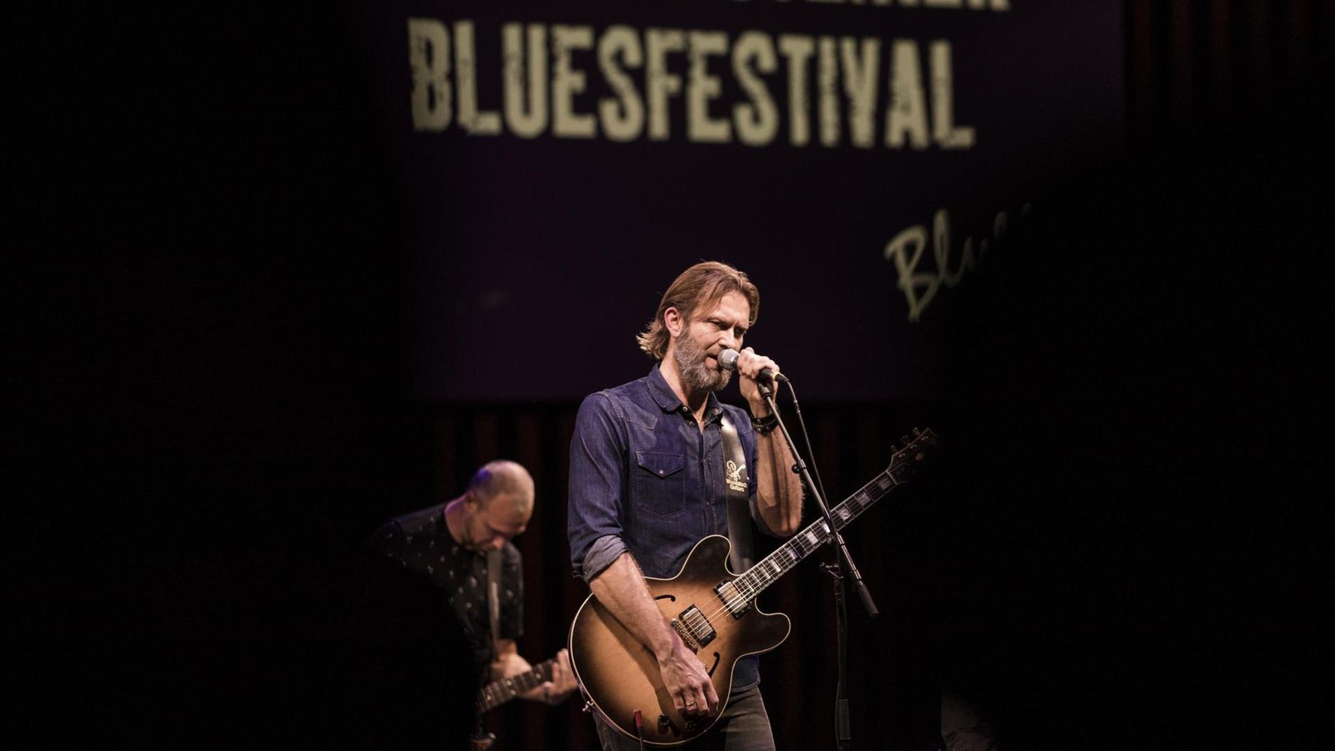 Ein Mann im blauen Hemd steht mit einer Gitarre in der Hand vor einem Mikrofon. Dahinter steht ein weiterer Musiker auf der Bühne. Ein Mann im blauen Hemd steht mit einer Gitarre in der Hand vor einem Mikrofon. Dahinter steht ein weiterer Musiker auf der Bühne.