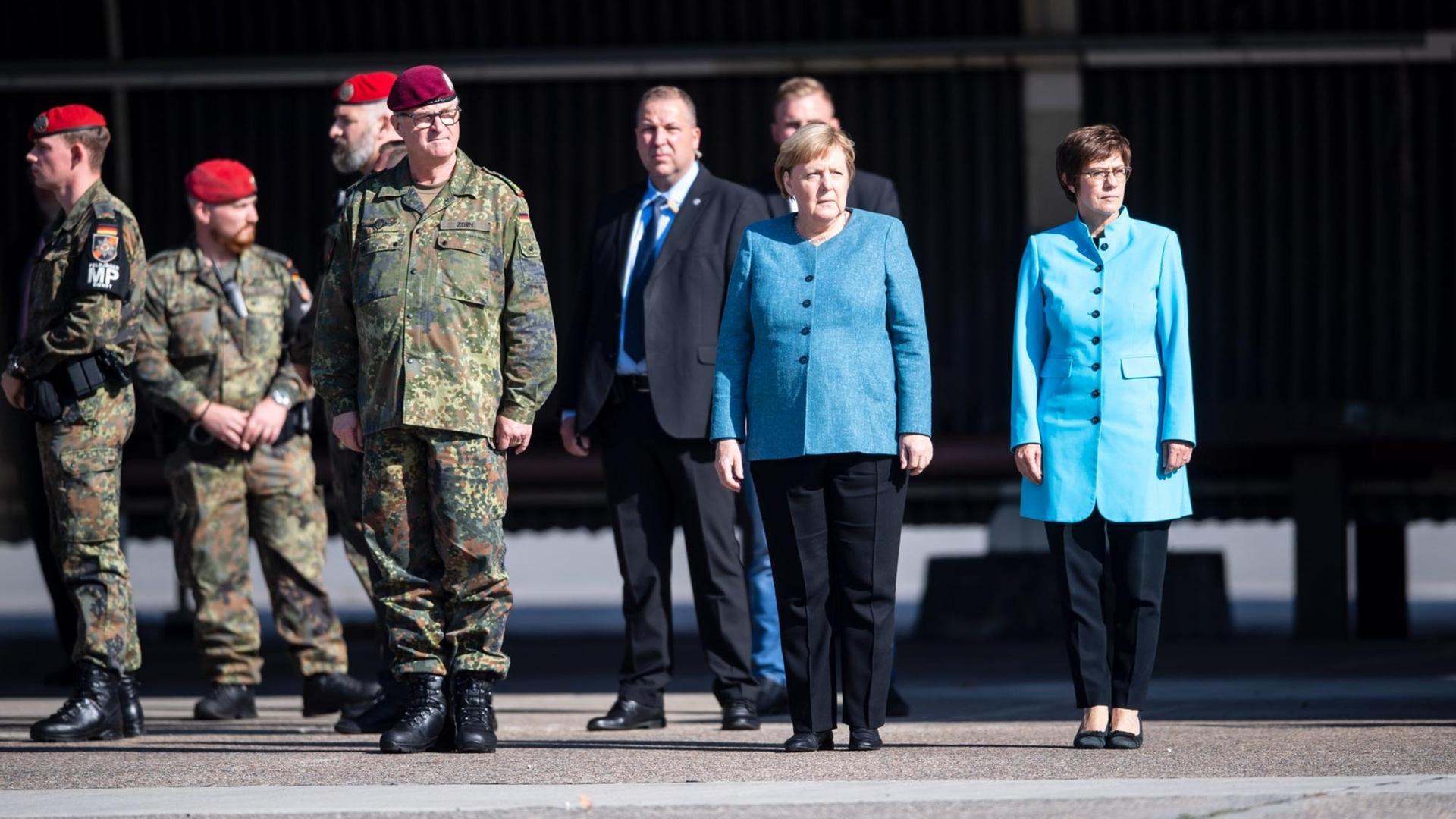 Die Bundeskanzlerin und Frau Kramp-Karrenbauer stehen neben mehreren Soldaten und anderen Wüdenträgern. Beide tragen mittelblaue Oberteile.