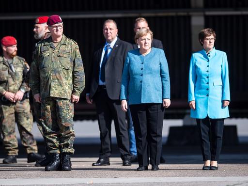 Die Bundeskanzlerin und Frau Kramp-Karrenbauer stehen neben mehreren Soldaten und anderen Wüdenträgern. Beide tragen mittelblaue Oberteile.