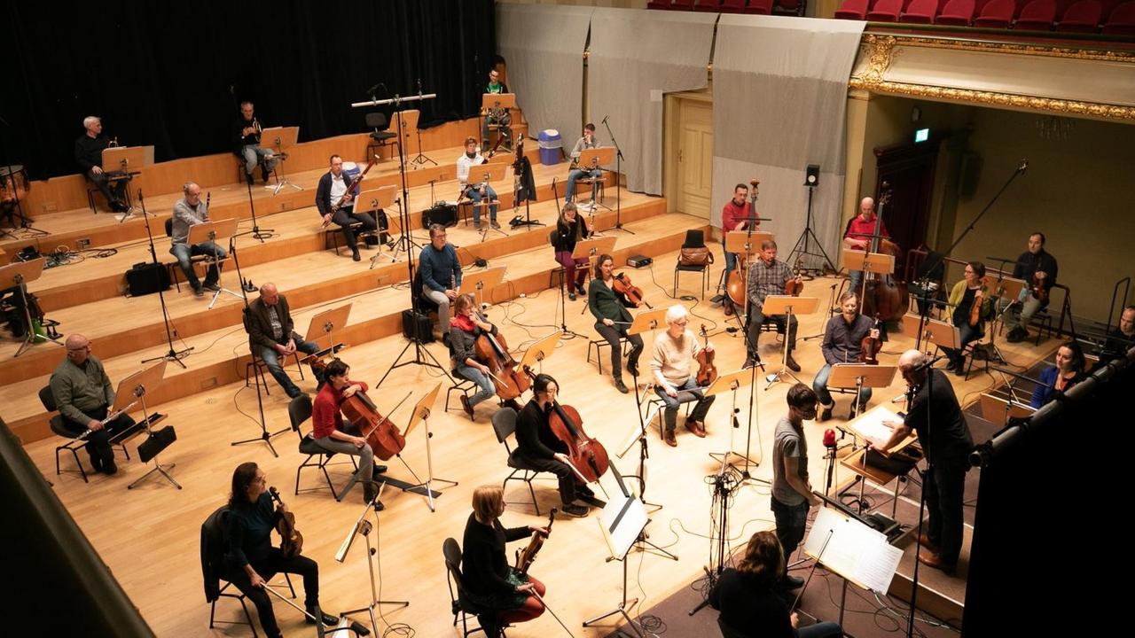 Ein Orchester sitzt mit großen Abständen auf einer hell beleuchteten Bühne und spielt. Ein Orchester sitzt mit großen Abständen auf einer hell beleuchteten Bühne und spielt.