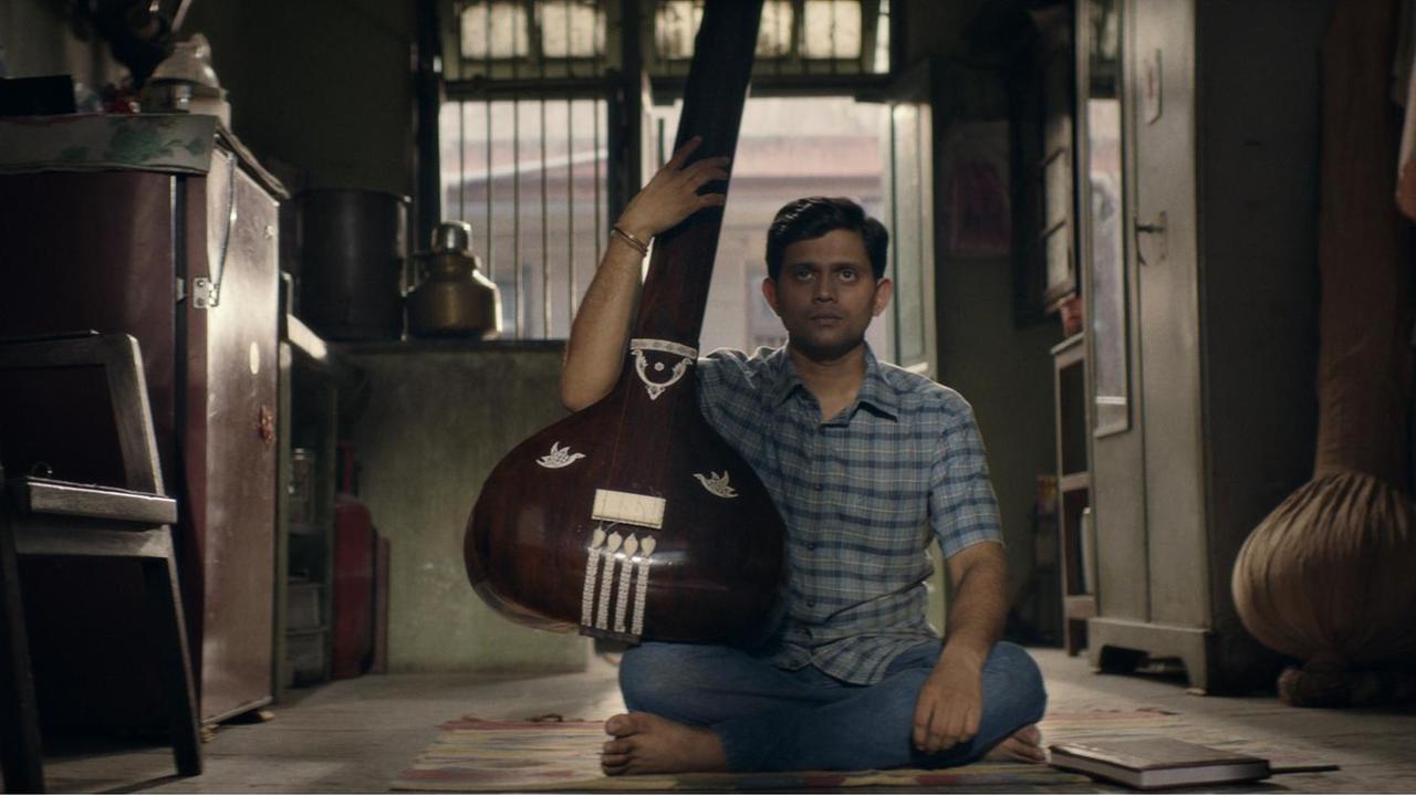 Filmszene aus "Der Schüler". Aditya Modak, in der Rolle des Sharad Nerulkar, sitzt mit einem traditionellen indischen Zupfinstrument auf dem Boden. Filmszene aus "Der Schüler". Aditya Modak, in der Rolle des Sharad Nerulkar, sitzt mit einem traditionellen indischen Zupfinstrument auf dem Boden.