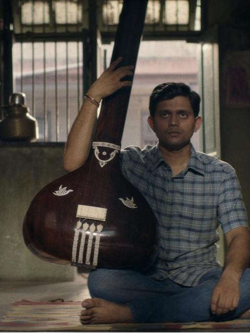 Filmszene aus "Der Schüler". Aditya Modak, in der Rolle des Sharad Nerulkar, sitzt mit einem traditionellen indischen Zupfinstrument auf dem Boden.