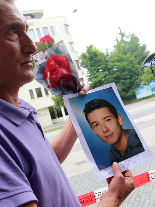 Naim Zabergja steht mit einem Foto seines getöteten Sohnes Dijamant am 23.07.2016 vor Medienvertretern am Olympia-Einkaufszentrum in München (Bayern), das die Polizei nach einer Schießerei mit Toten und Verletzten am Vortag abgesperrt hat.