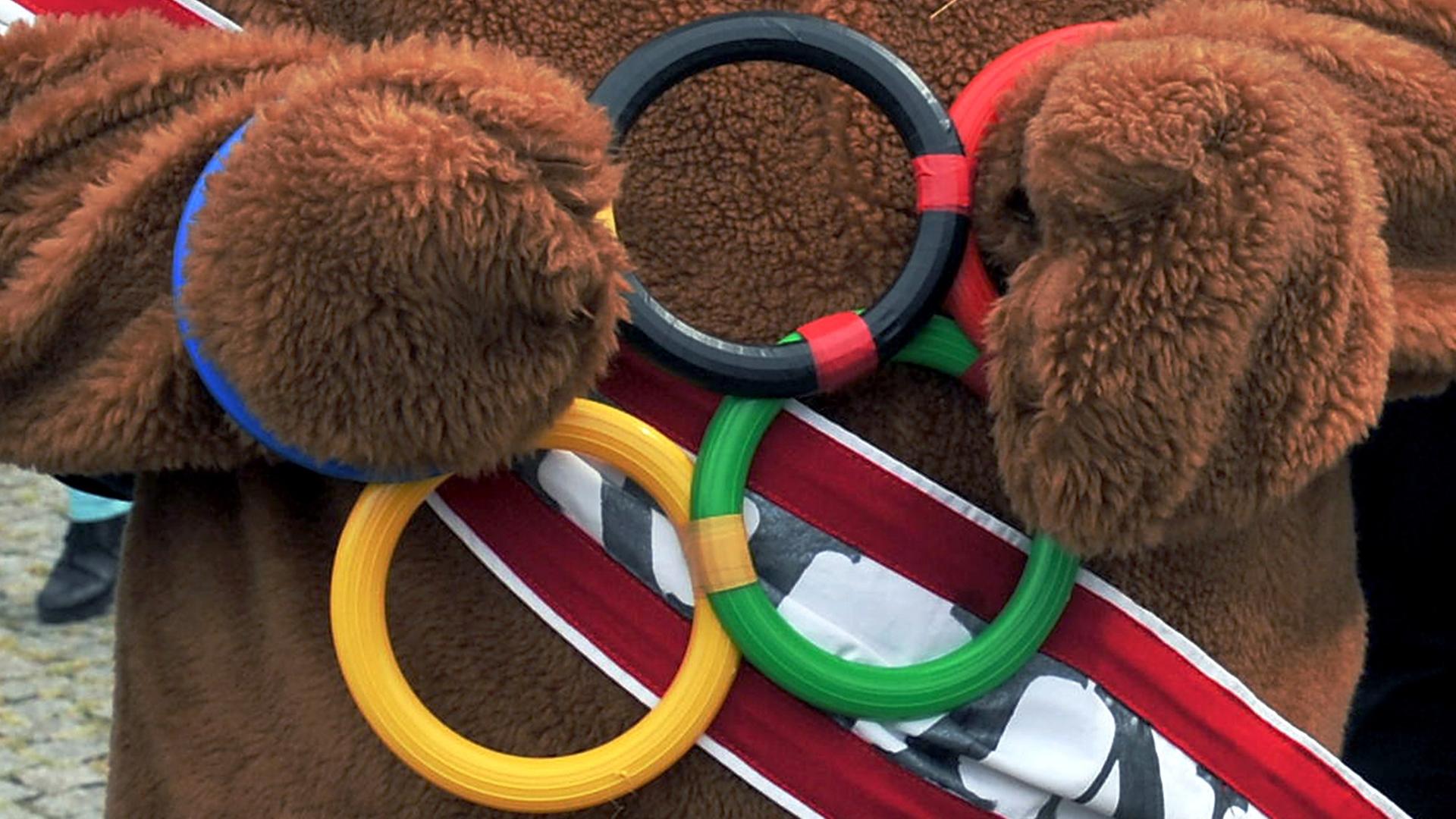Ein als Bär verkleideter Mann streckt die Hände durch symbolische Olympische Ringe. Ein als Bär verkleideter Mann streckt die Hände durch symbolische Olympische Ringe.