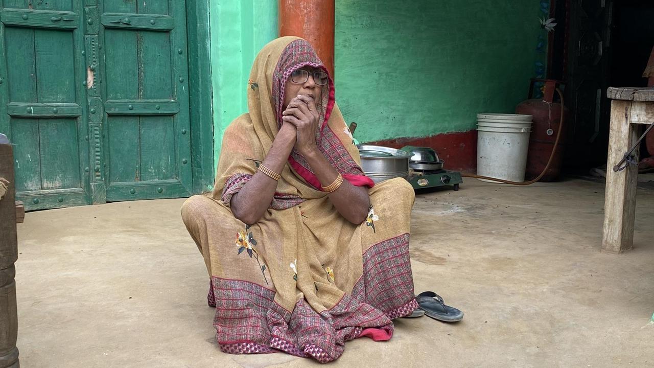Eine ältere Frau sitzt mit einem langen beigen  Gewand und bedecktem Kopf auf dem Lehmboden ihres Hauses. Und faltet die Hände  vor dem Gesicht.
