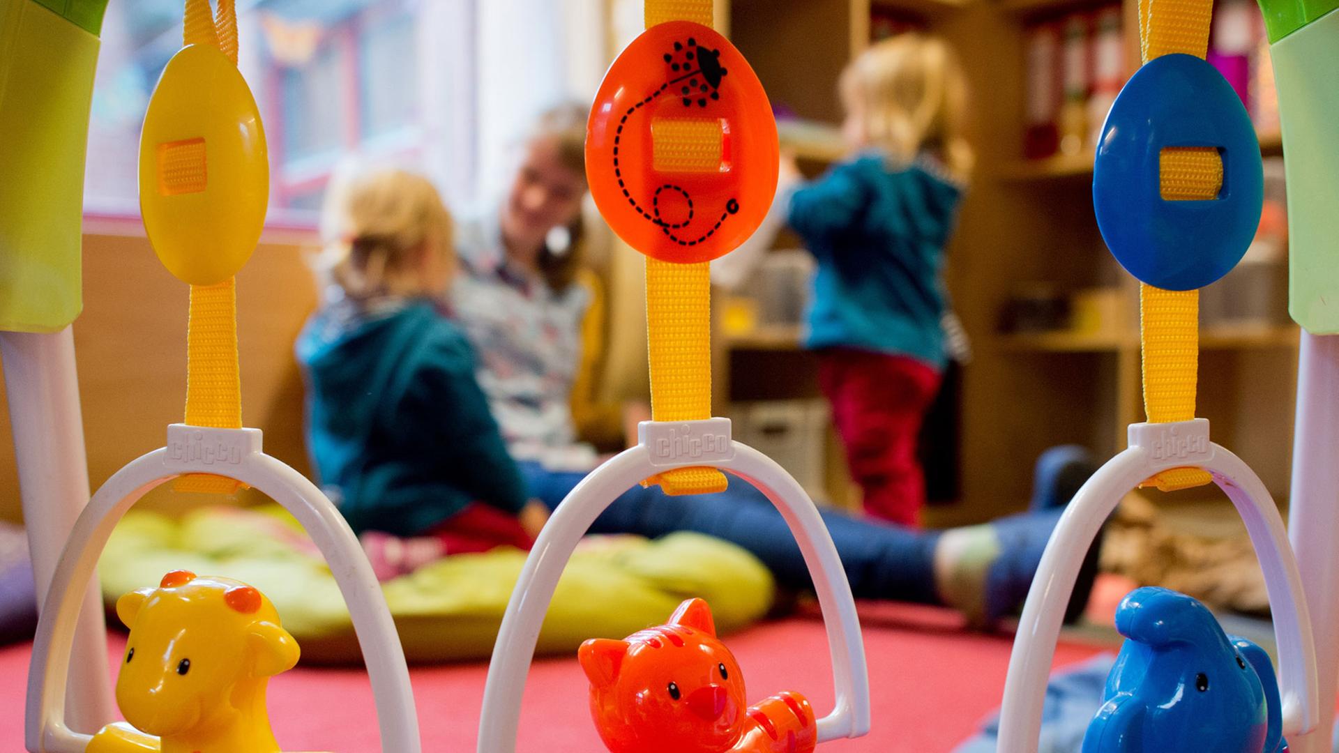Kinderspielzeug hängt an einem Rahmen. Im Hintergrund spielt eine Erzieherin mit zwei kleinen Kindern Kinderspielzeug hängt an einem Rahmen. Im Hintergrund spielt eine Erzieherin mit zwei kleinen Kindern