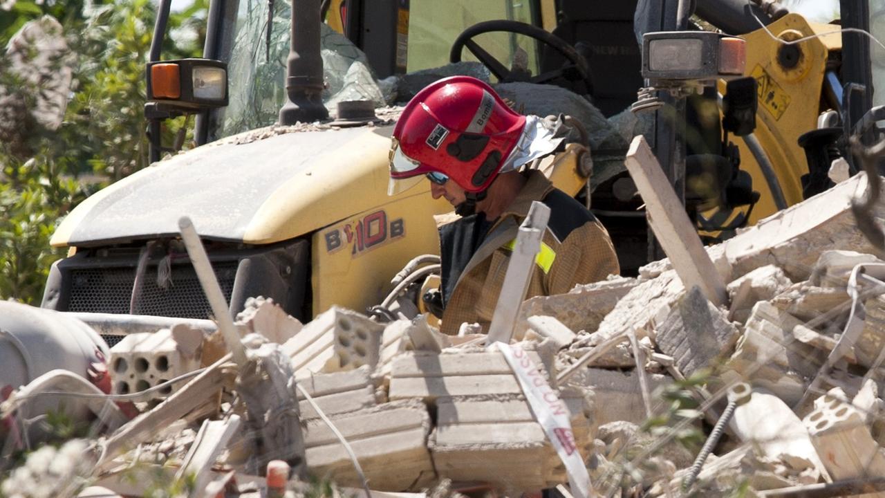 Der Ermittler mit rotem Helm und Sonnenbrille steht vor einem gelben Traktor und hinter einem Trümmerhaufen. Der Ermittler mit rotem Helm und Sonnenbrille steht vor einem gelben Traktor und hinter einem Trümmerhaufen.