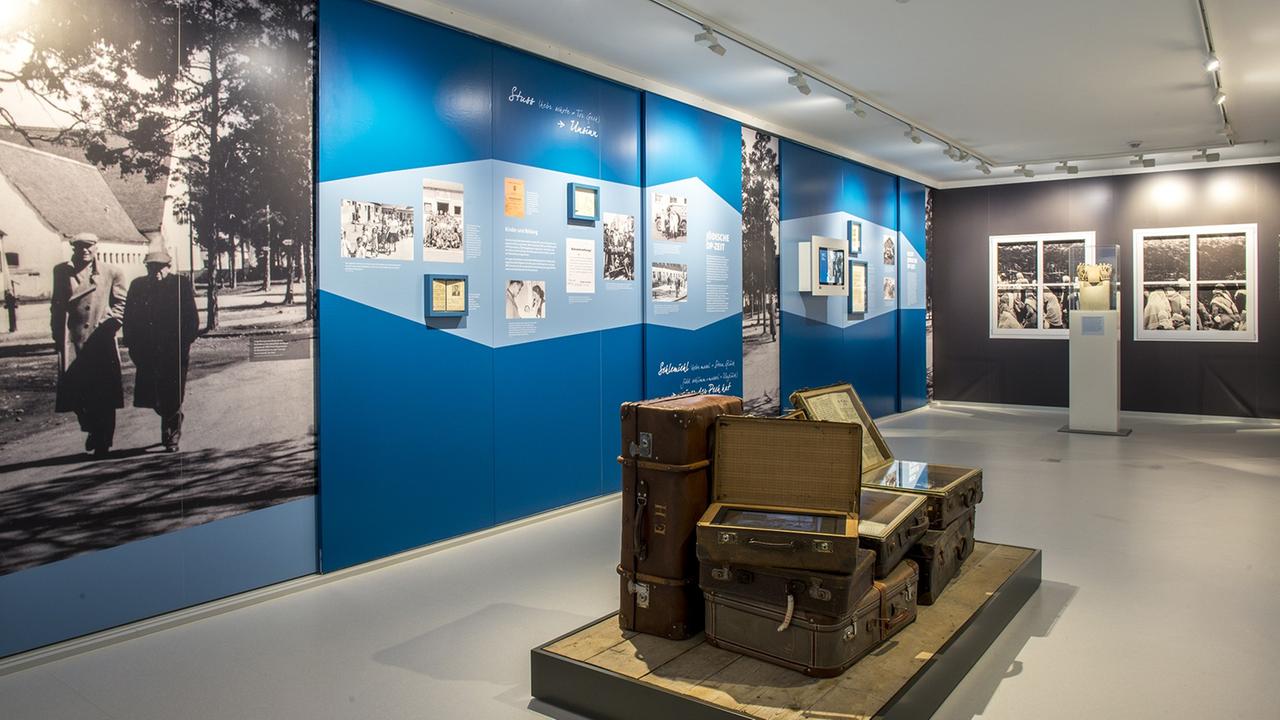 Blick in die Ausstellung des Erinnerungsorts Badehaus im ehemaligen DP-Lager Föhrenwald und heutigen Waldram in Oberbayern: mehrere Koffer umgeben von Bildwänden und weiteren Ausstellungsstücken. Blick in die Ausstellung des Erinnerungsorts Badehaus im ehemaligen DP-Lager Föhrenwald und heutigen Waldram in Oberbayern: mehrere Koffer umgeben von Bildwänden und weiteren Ausstellungsstücken.