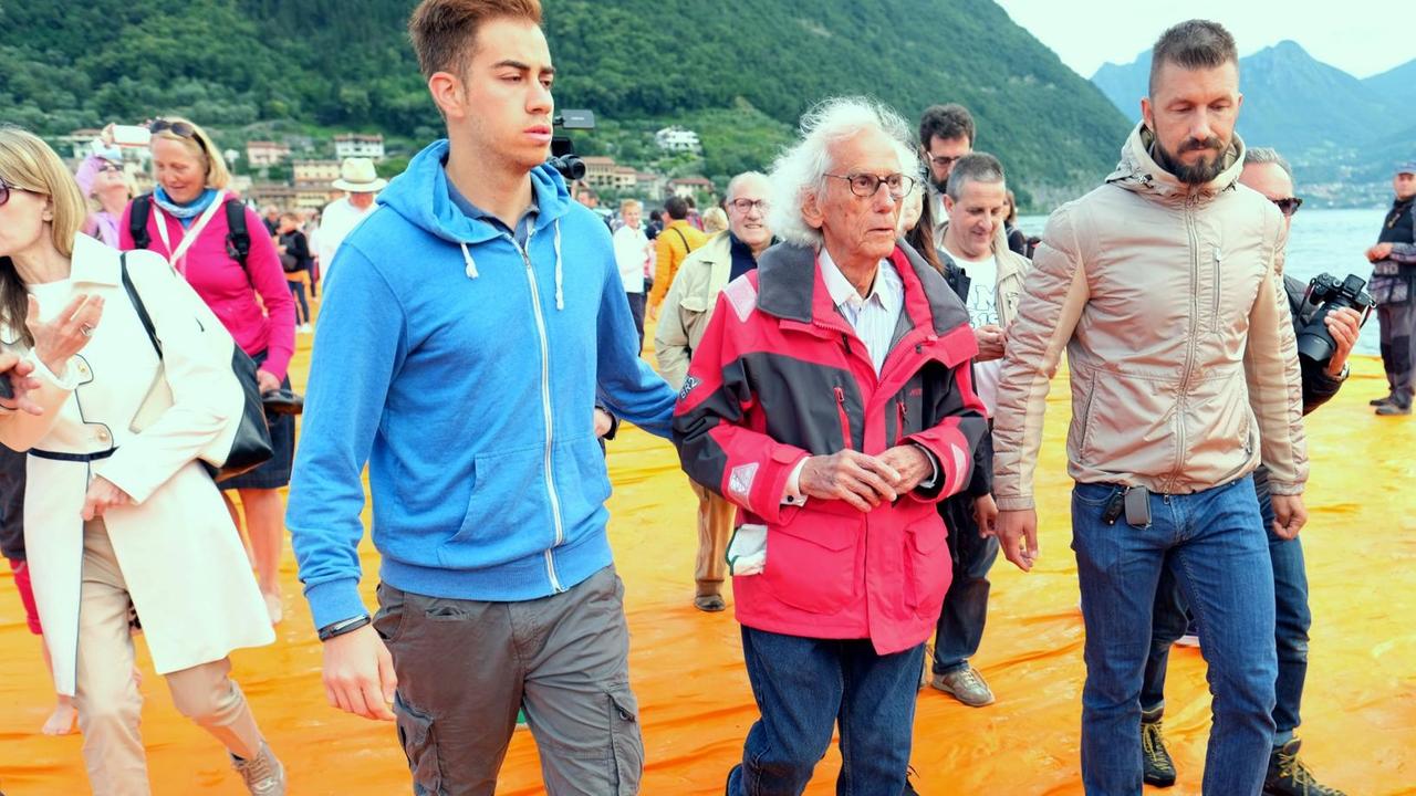 Künstler Christo (M) wird am 18.06.2016 vor Paratico auf dem Lago d'Iseo in Italien von Begleitern durch die Besuchermassen über die schwimmendenen Stege mit orangefarbenen Stoff seines Projekts "The Floating Piers" geführt. Künstler Christo (M) wird am 18.06.2016 vor Paratico auf dem Lago d'Iseo in Italien von Begleitern durch die Besuchermassen über die schwimmendenen Stege mit orangefarbenen Stoff seines Projekts "The Floating Piers" geführt.
