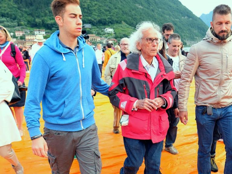 Künstler Christo (M) wird am 18.06.2016 vor Paratico auf dem Lago d'Iseo in Italien von Begleitern durch die Besuchermassen über die schwimmendenen Stege mit orangefarbenen Stoff seines Projekts "The Floating Piers" geführt.