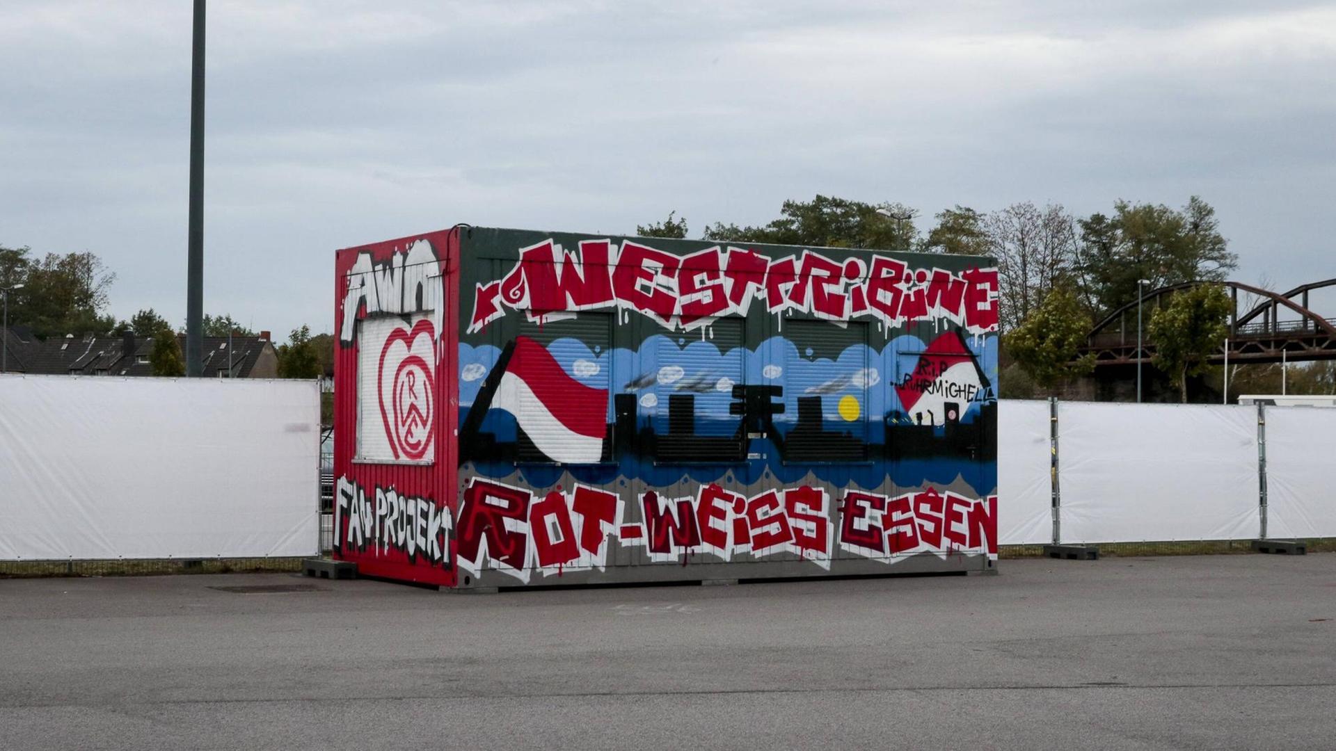 Ein mit Graffiti besprühter Container steht vor einem Fußballstadion.