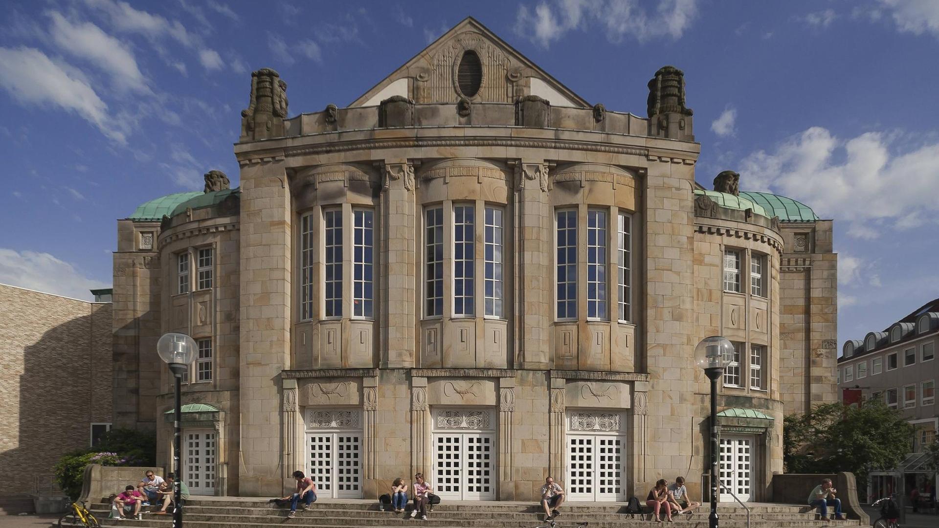 Vor einem historischen Bau mit großen Fenstern und Schmuck an der Fassade sitzen Menschen auf den wenigen Eingangsstufen, die sich in der Vorderfront des Gebäudes zeigen. Vor einem historischen Bau mit großen Fenstern und Schmuck an der Fassade sitzen Menschen auf den wenigen Eingangsstufen, die sich in der Vorderfront des Gebäudes zeigen.