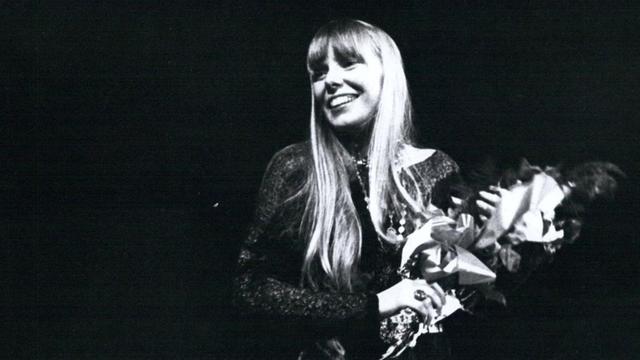 Joni Mitchell in einer Schwarzaufnahme: Sie steht auf einer Bühne und hält einen Strauß Blumen in der Hand, während sie fröhlich ins Publikum lacht.