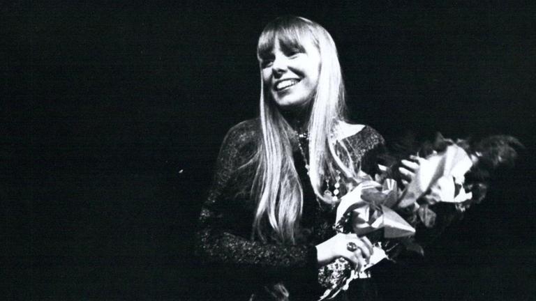 Joni Mitchell in einer Schwarzaufnahme: Sie steht auf einer Bühne und hält einen Strauß Blumen in der Hand, während sie fröhlich ins Publikum lacht. Joni Mitchell in einer Schwarzaufnahme: Sie steht auf einer Bühne und hält einen Strauß Blumen in der Hand, während sie fröhlich ins Publikum lacht.