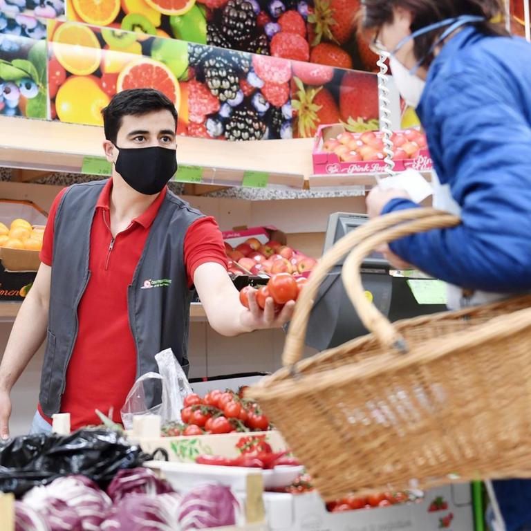 22.04.2020, Bayern, Rosenheim: Ein Mitarbeiter eines Lebensmittelgeschäfts bedient in der Gemüseabteilung eine Kundin und trägt eine Maske als Mund- und Nasenschutz.