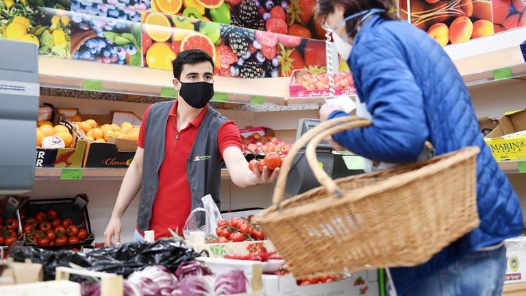 22.04.2020, Bayern, Rosenheim: Ein Mitarbeiter eines Lebensmittelgeschäfts bedient in der Gemüseabteilung eine Kundin und trägt eine Maske als Mund- und Nasenschutz.
