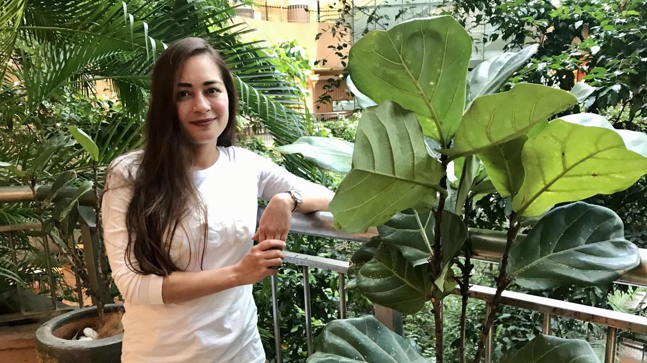 Eine junge Frau mit langen glatten dunkelbraunen Haaren, Jeans und weißem T-Shirt steht inmitten von Grünpflanzen und stützt sich auf ein Geländer. Eine junge Frau mit langen glatten dunkelbraunen Haaren, Jeans und weißem T-Shirt steht inmitten von Grünpflanzen und stützt sich auf ein Geländer.