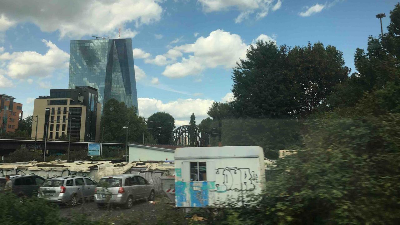 Das Gebäude der Europäischen Zentralbank in Frankfurt am Main aus dem Zug heraus fotografiert. Im Vordergrund parkende Autos und ein Bauwagen. 