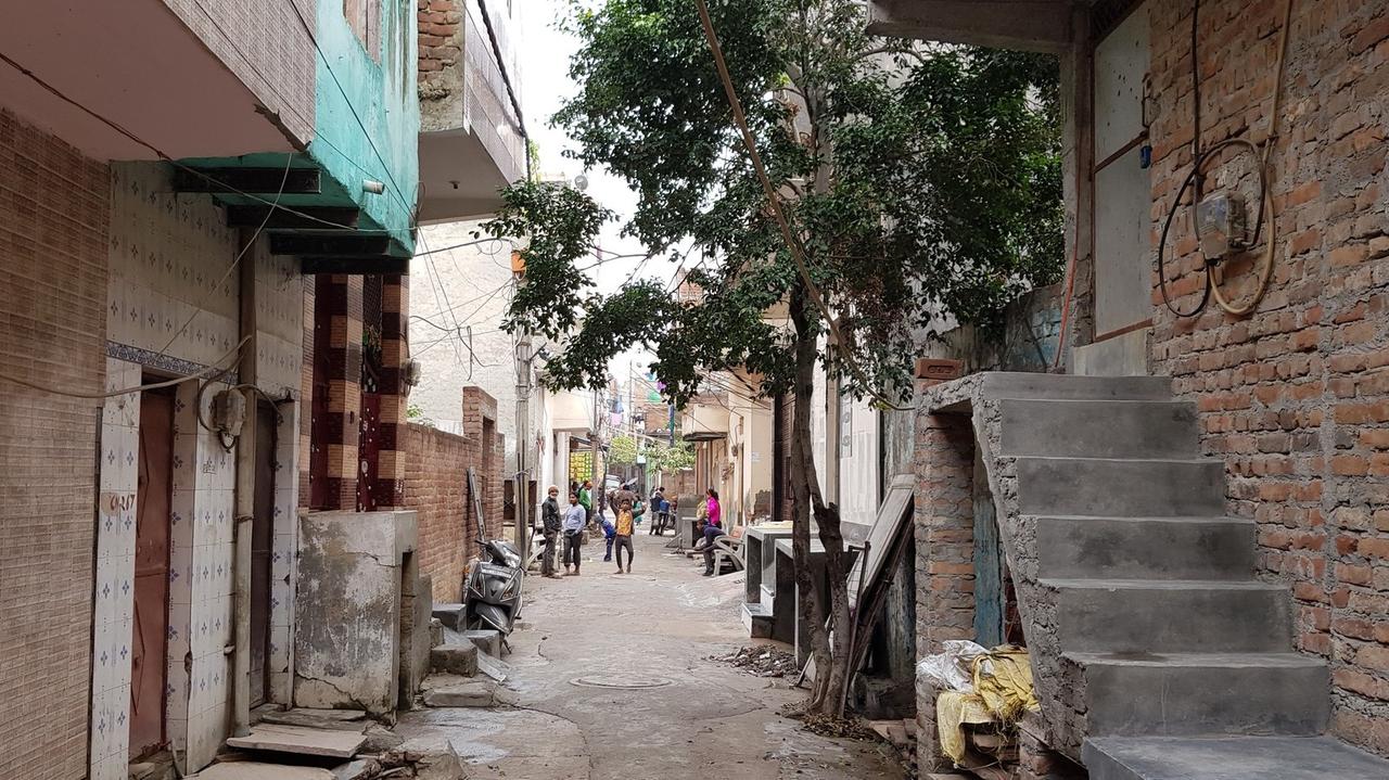Blick in eine enge Gasse in Shiv Vihar, einem Stadtteil der indischen Hauptstadt Neu-Delhi, im Vordergrund ein schlanker Baum und eine Treppe, die an einer unverputzten Hauswand hoch führt, hinten Menschen in gelben, grünen, rosa Hemden