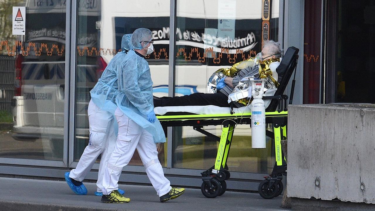 Eine alte Frau liegt mit Beatmungsgerät auf einer Liege und wird von zwei Personen in ein Krankenhaus gerollt. Eine alte Frau liegt mit Beatmungsgerät auf einer Liege und wird von zwei Personen in ein Krankenhaus gerollt.