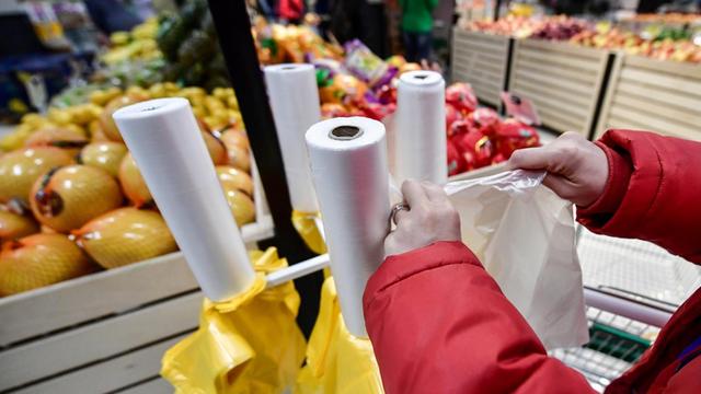 In einem Supermarkt reißt ein Kunde eine Plastiktüte von der Rolle, um Obst und Gemüse einzupacken. Im Hintergrund sieht man das Angebot in der Obst- und Gemüseabteilung: Melonen, Äpfel und Orangen.
