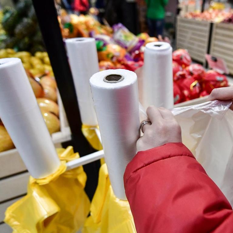 In einem Supermarkt reißt ein Kunde eine Plastiktüte von der Rolle, um Obst und Gemüse einzupacken. Im Hintergrund sieht man das Angebot in der Obst- und Gemüseabteilung: Melonen, Äpfel und Orangen.