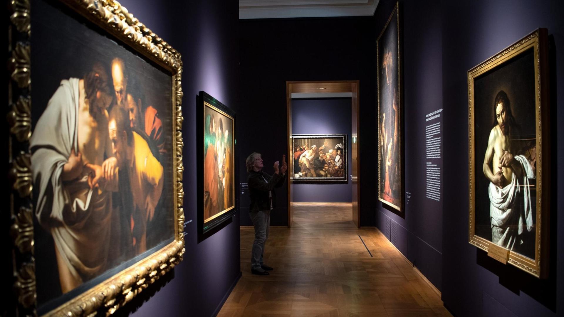 Ein Gang mit dunkellila Wänden und großflächigen Gemälden in der Ausstellung "Utrecht, Caravaggio und Europa", eine Frau fotografiert eines der Gemälde.