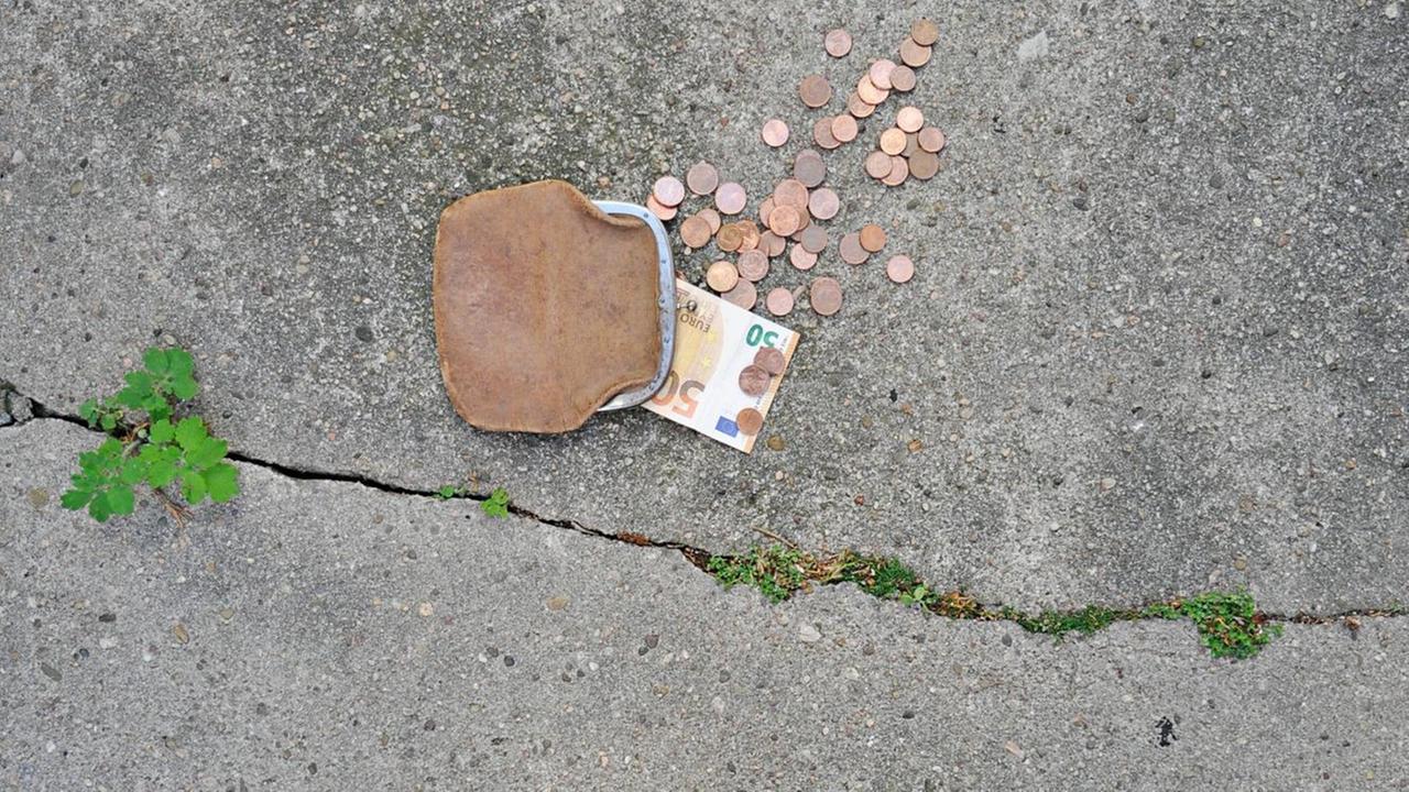Eine braune Ledergeldbörse mit Schnappverschluss liegt auf dem Boden. Ein Fünfzig-Euro-Schein ragt heraus und mehrere Cent-Münzen liegen verstreut. Eine braune Ledergeldbörse mit Schnappverschluss liegt auf dem Boden. Ein Fünfzig-Euro-Schein ragt heraus und mehrere Cent-Münzen liegen verstreut.