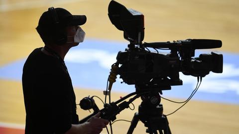 Ein Kameramann steht mit Mundschutz an seiner TV-Kamera bei einem Basketball vor leeren Publikumsplätzen.