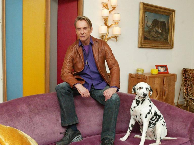 Porträt von Wolfgang Joop in seiner Villa Wunderkind in Potsdam. Er sitzt auf der Lehne eines rosa farbenen Sofa zusammen mit seinem Dalmatiner Hund.