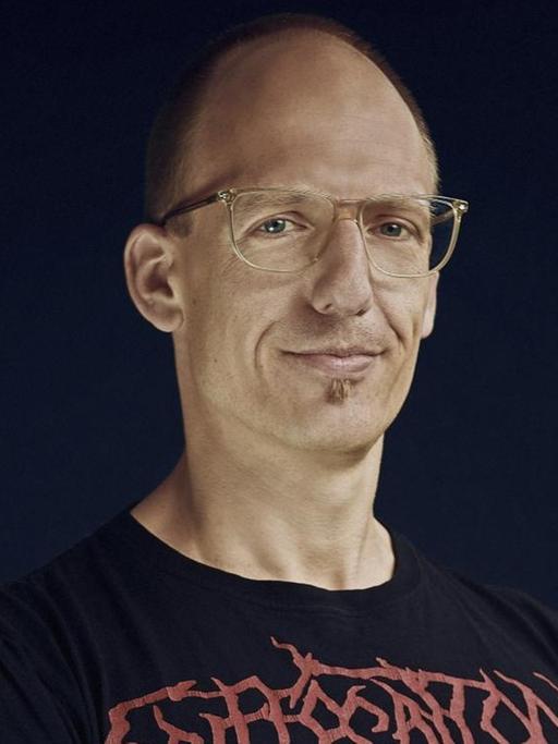 Jörg Scheller trägt eine Brille und ein schwarzes T-Shirt mit dem Namensschriftzug einer Heavy Metal Band