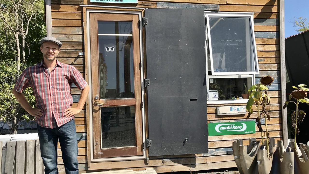 Ein freundlicher junger Mann mit Karohemd, Jeans und Käppi steht vor einer kleinen Holzhütte. Ein freundlicher junger Mann mit Karohemd, Jeans und Käppi steht vor einer kleinen Holzhütte.