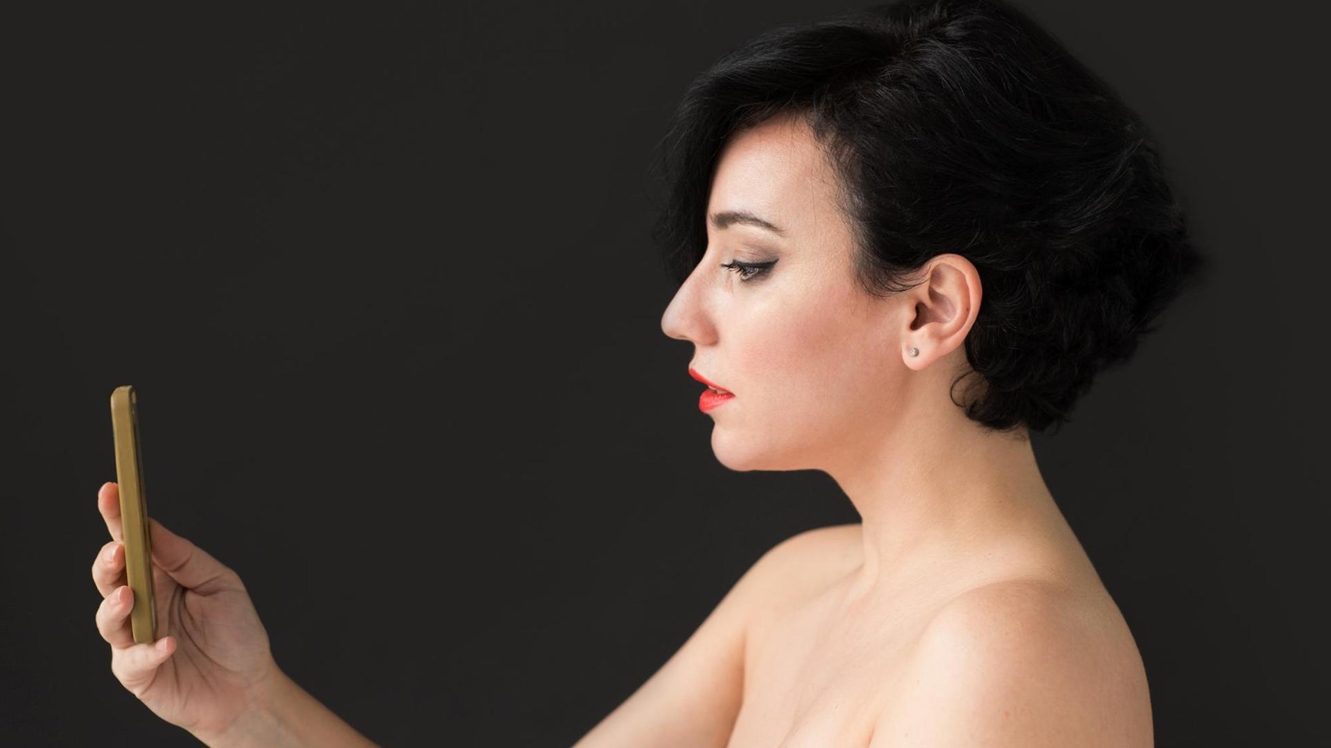Ein Frau mit nackten Schultern macht ein Selfie von sich. Der Hintergrund ist schwarz. Ein Frau mit nackten Schultern macht ein Selfie von sich. Der Hintergrund ist schwarz.
