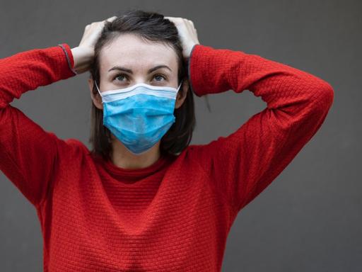 Eine Frau mit Mund- und Nasenschutzmaske fasst sich mit beiden Händen an den Kopf.