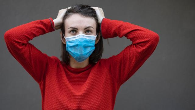 Eine Frau mit Mund- und Nasenschutzmaske fasst sich mit beiden Händen an den Kopf.