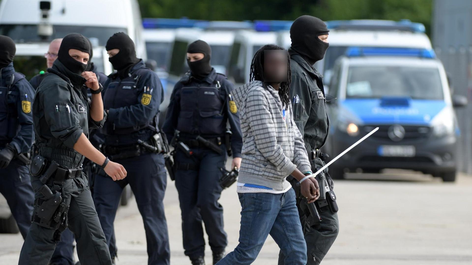 Ein maskierter Polizist führt einen gefesselten Mann ab. Das Gesicht des Schwarzafrikaners ist verpixelt. Dahinter weitere maskierte Beamte und Polizeiwagen. Ein maskierter Polizist führt einen gefesselten Mann ab. Das Gesicht des Schwarzafrikaners ist verpixelt. Dahinter weitere maskierte Beamte und Polizeiwagen.