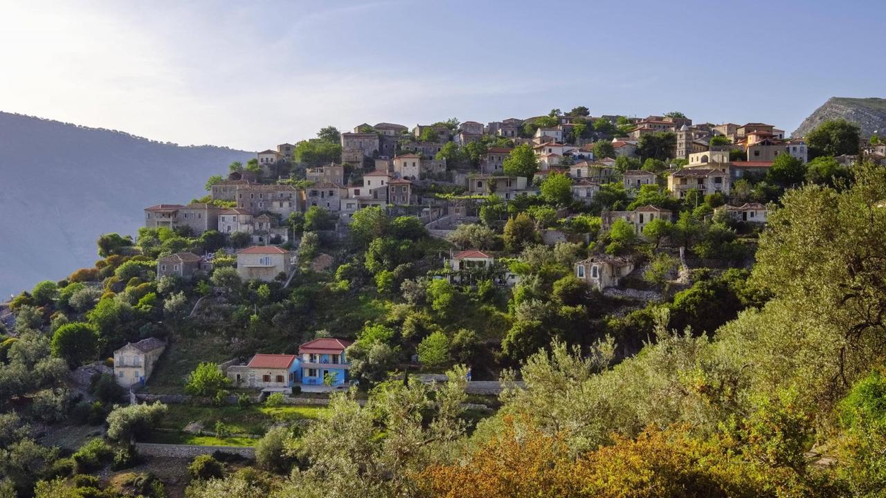 Das Bergdorf Qeparo an der Albanischen Riviera: Über einen mit Bäumen bewachsenen Hügel verstreut stehen Häuser, im Hintergrund sind weitere Erhebungen zu sehen.