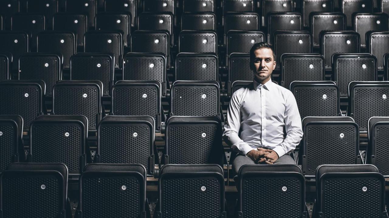 Der Stuttgarter Comedian Özcan Cosar sitzt alleine in einem Konzertsaal mit schwarzen Klappstühlen.