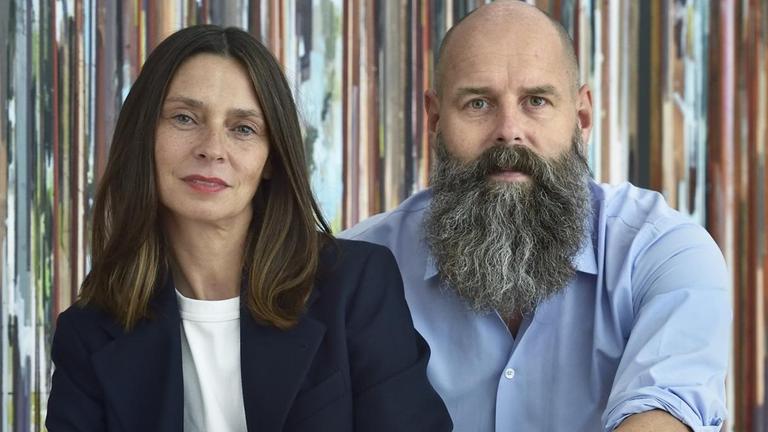 Porträtfoto von Silke und Holger Friedrich, die nebeneinander vor einer bunt gestreiften Wand sitzen. Porträtfoto von Silke und Holger Friedrich, die nebeneinander vor einer bunt gestreiften Wand sitzen.